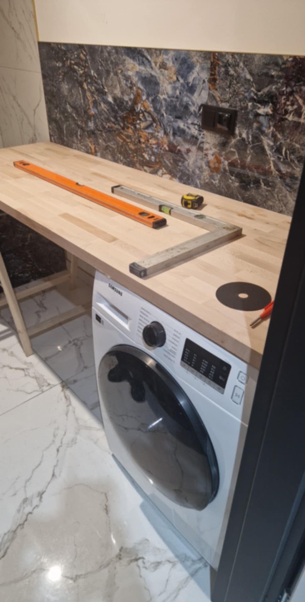 Blat z jasnego drewna nad pralką, poziomica, kątownik i miara zwinięta, przygotowane do montażu; tło stanowi marmurowa ściana i podłoga.