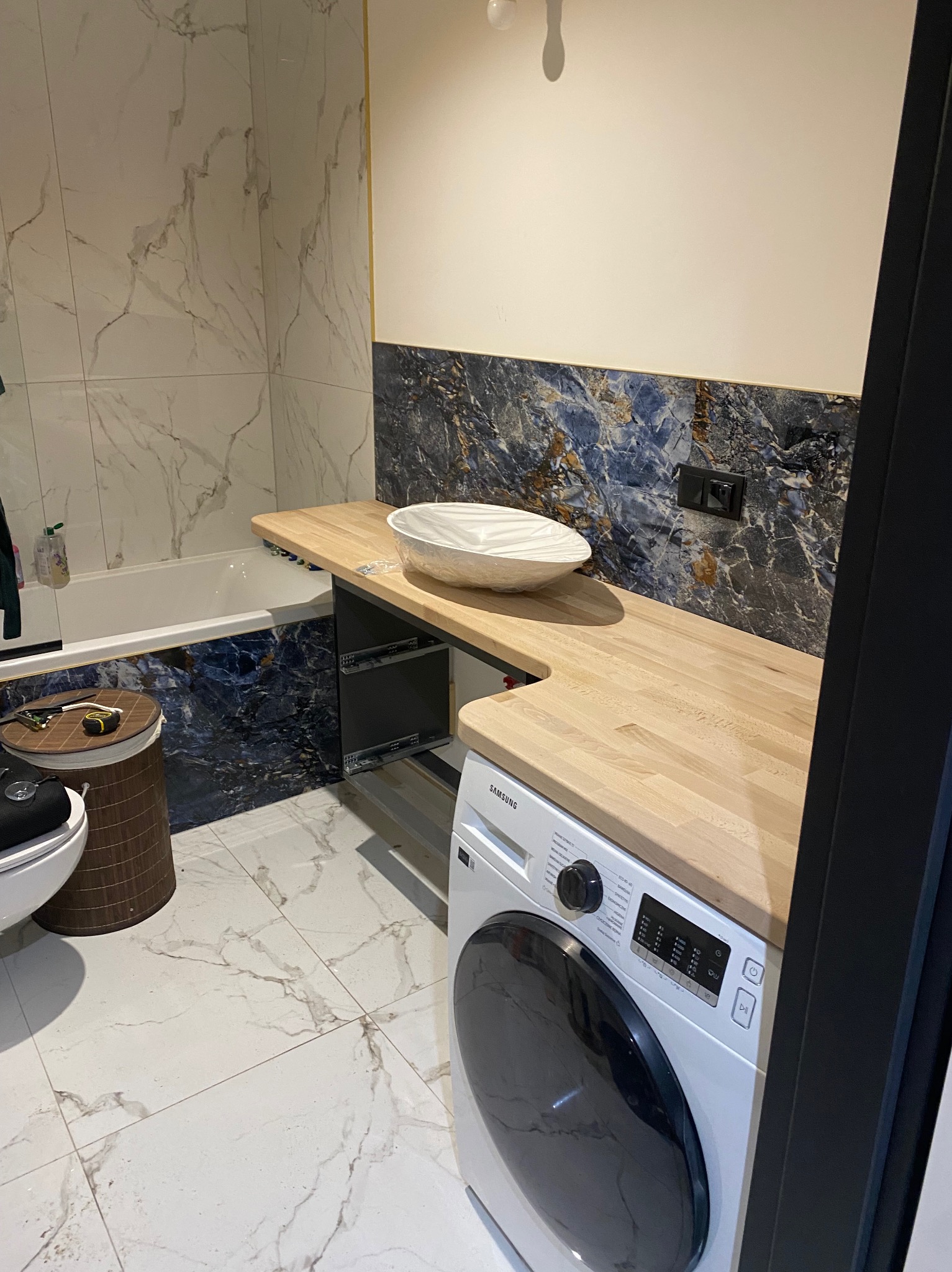 Łazienka w trakcie remontu: blat z jasnego drewna zamontowany nad pralką Samsung, umywalka tymczasowo zabezpieczona folią, marmurowe płytki na ścianach i podłodze, widoczne elementy montażowe szuflad.