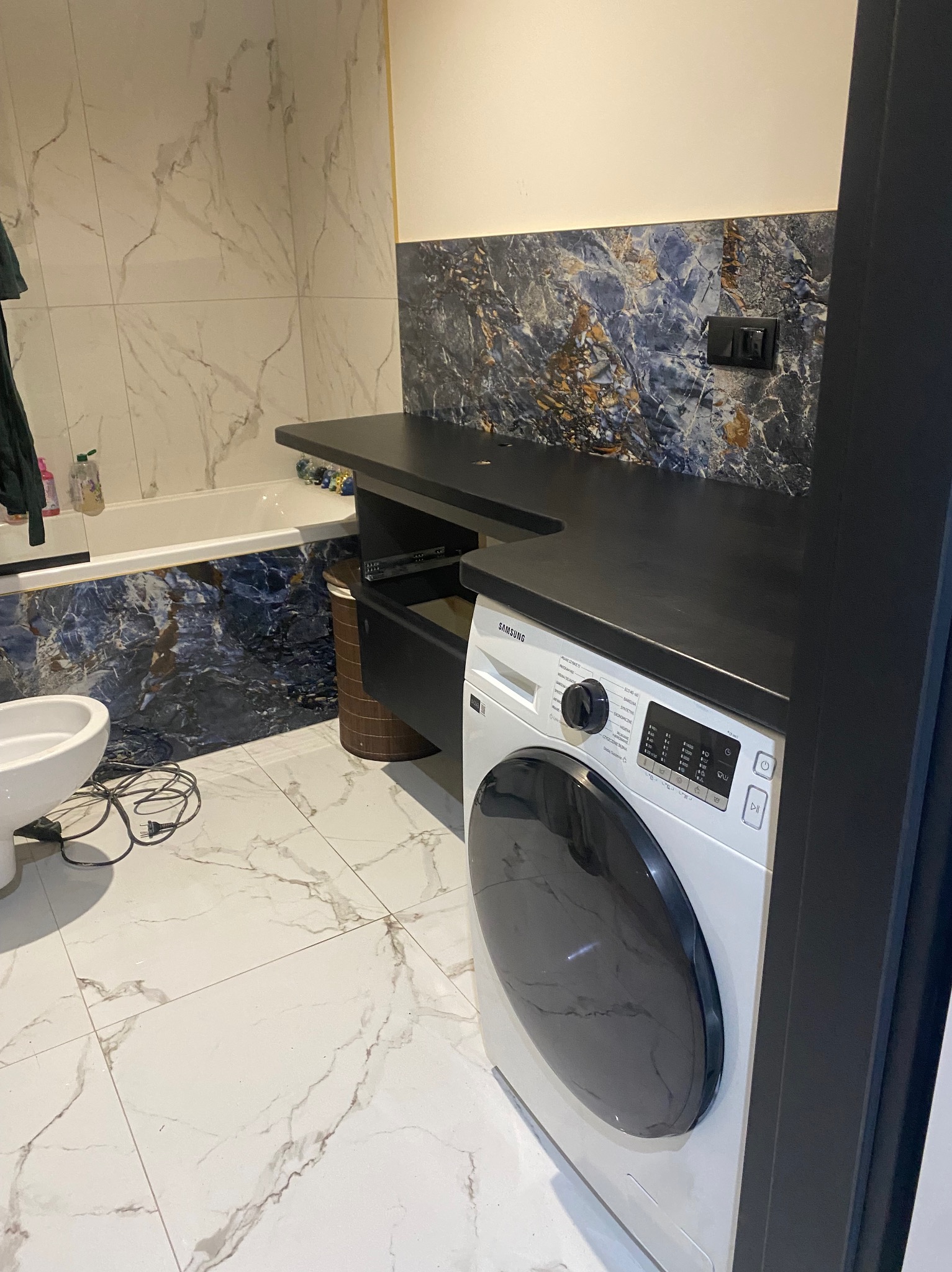 Wykończona łazienka z pralką Samsung, białą armaturą i marmurowymi płytkami na ścianach i podłodze, widoczne czarne blaty i ciemnoniebieskie płytki dekoracyjne.