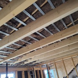 MULTI ENERGA SPÓŁKA Z OGRANICZONĄ ODPOWIEDZIALNOŚCIĄ - Wnętrze poddasza w trakcie ocieplania: widoczne drewniane belki stropowe, metalowe profile pod płyty gipsowe oraz brązowa wełna mineralna między nimi.