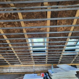 MULTI ENERGA SPÓŁKA Z OGRANICZONĄ ODPOWIEDZIALNOŚCIĄ - Poddasze w trakcie ocieplania wełną mineralną, widoczne metalowe profile mocujące i okna dachowe, na dole składowane płyty izolacyjne i butelka wody.