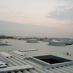 MULTI ENERGA SPÓŁKA Z OGRANICZONĄ ODPOWIEDZIALNOŚCIĄ - Pracownik w kasku i kamizelce odblaskowej na dachu przemysłowym z blachy trapezowej, widoczne otwory okienne, obróbki blacharskie i materiały budowlane w tle.