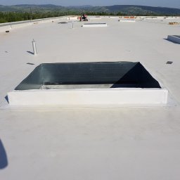 MULTI ENERGA SPÓŁKA Z OGRANICZONĄ ODPOWIEDZIALNOŚCIĄ - Biała, płaska powierzchnia dachu z zainstalowanymi świetlikami i kominkami wentylacyjnymi, w tle widoczni pracownicy i górzysty krajobraz.