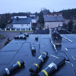 MULTI ENERGA SPÓŁKA Z OGRANICZONĄ ODPOWIEDZIALNOŚCIĄ - Bitumiczna papa w rolkach leży na płaskim dachu pokrytym czarnym materiałem izolacyjnym, widoczne uszczelnienia pianką montażową, w tle zabudowa jednorodzinna i las.