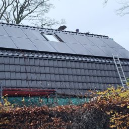 MULTI ENERGA SPÓŁKA Z OGRANICZONĄ ODPOWIEDZIALNOŚCIĄ - Montaż paneli fotowoltaicznych na dachu pokrytym ciemną dachówką, widoczna drabina oparta o dach i pracownik w niebieskiej czapce na szczycie.