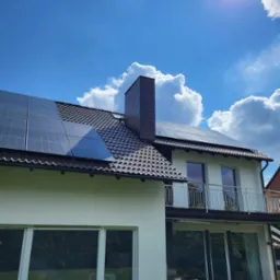 Dom z brązowym dachem i panelami słonecznymi w słoneczny dzień, widoczne białe ściany, balkon z białą balustradą i komin z ciemnej dachówki.