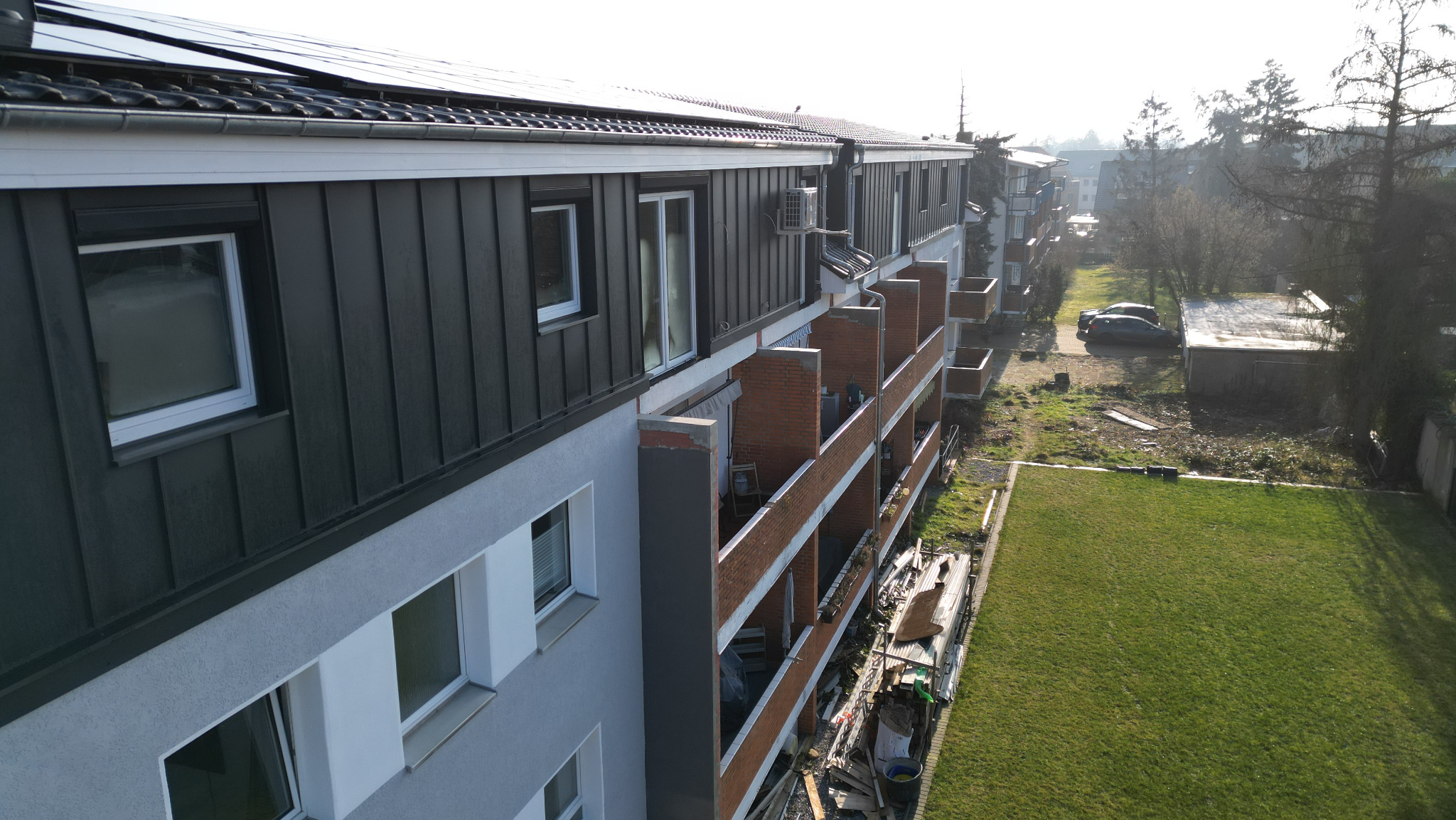 Wielopiętrowy budynek z panelami słonecznymi na dachu, widok z góry na grafitową blachę na elewacji i rynny, balkony z ceglaną balustradą.