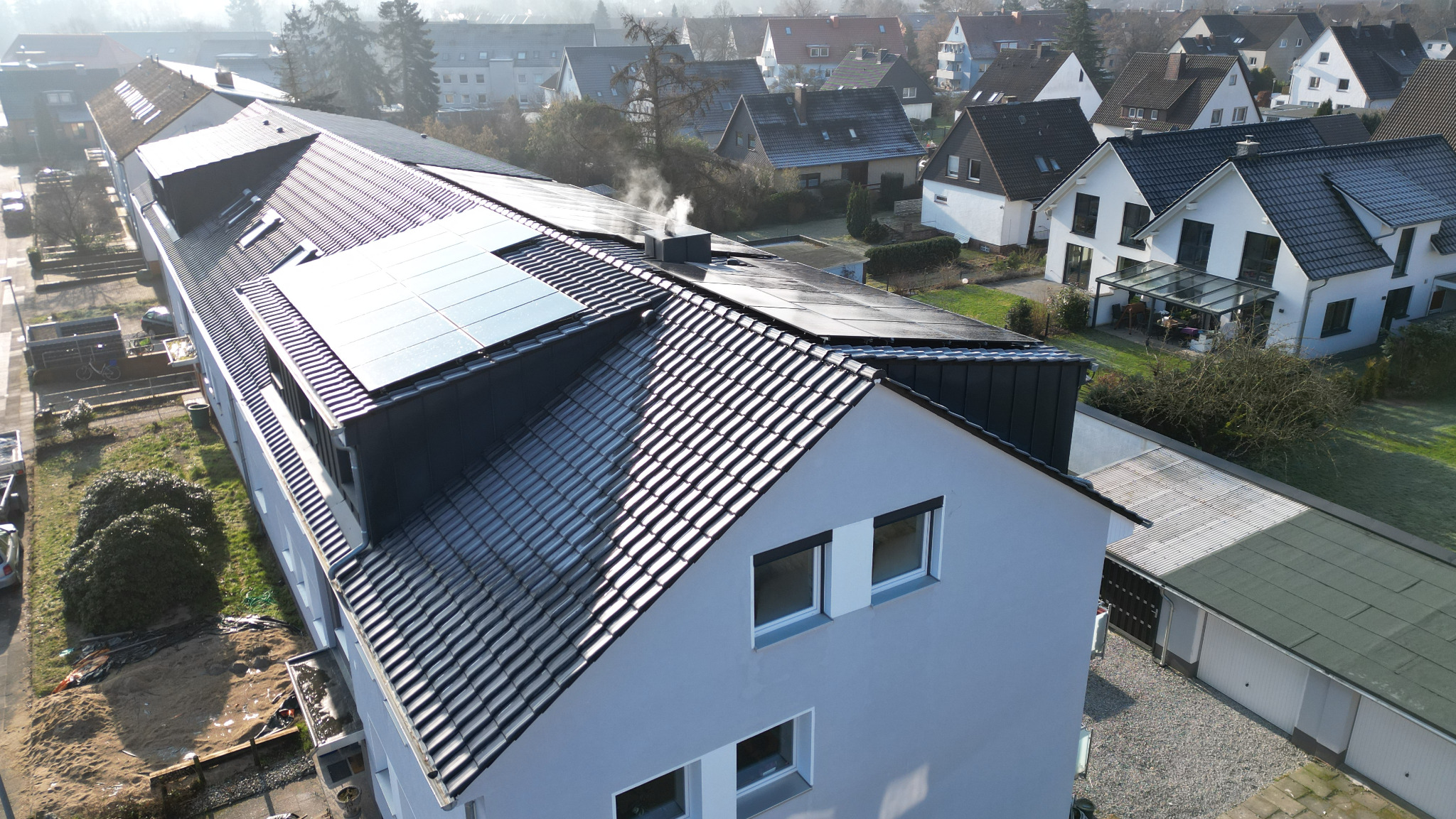 Widok z góry na budynek wielorodzinny z ciemną dachówką i panelami słonecznymi, z komina unosi się dym, w tle zabudowa jednorodzinna.