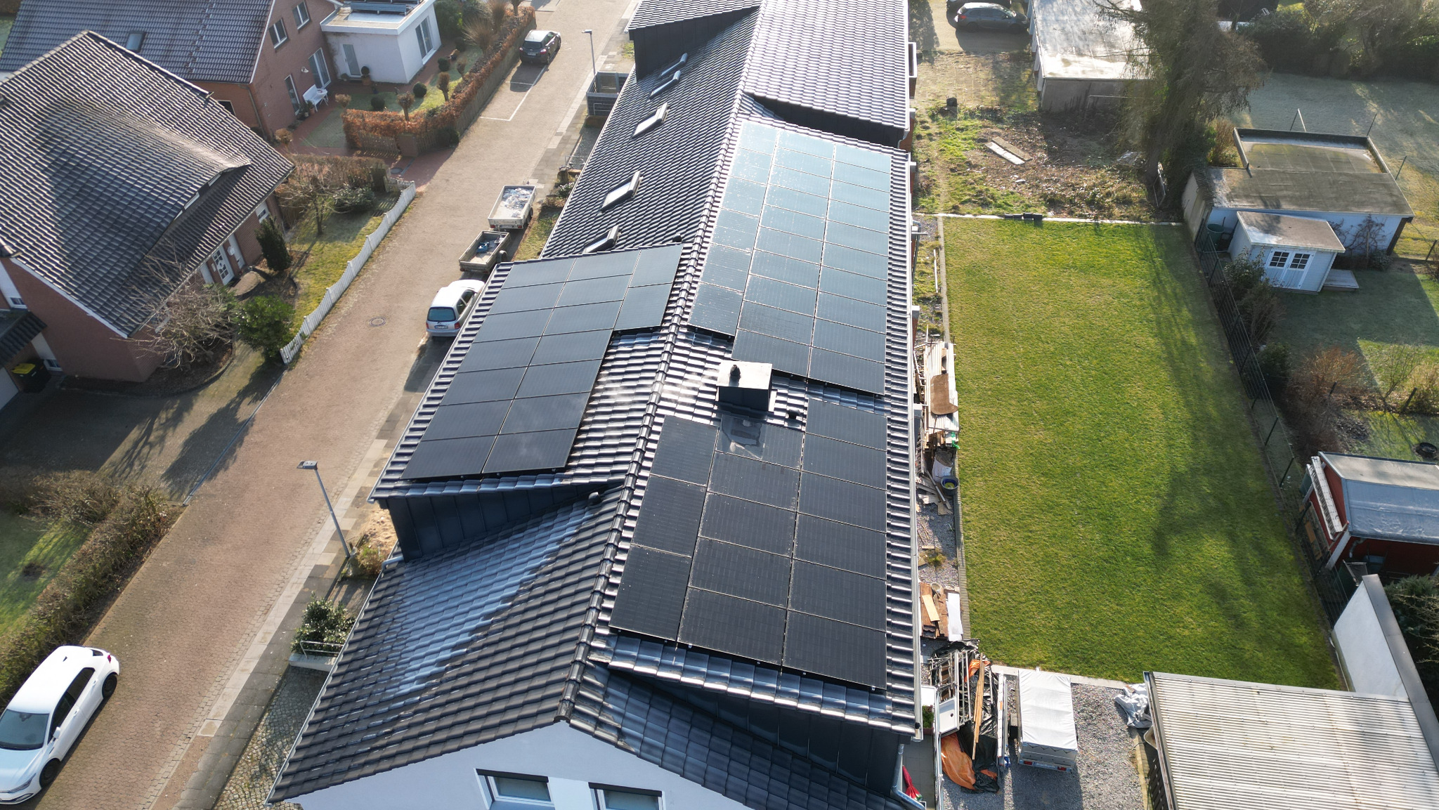 Widok z góry na dom z ciemnoszarą dachówką i panelami słonecznymi, z widocznym kominem i elementami instalacji na dachu, otoczony zielenią i zabudową mieszkalną.