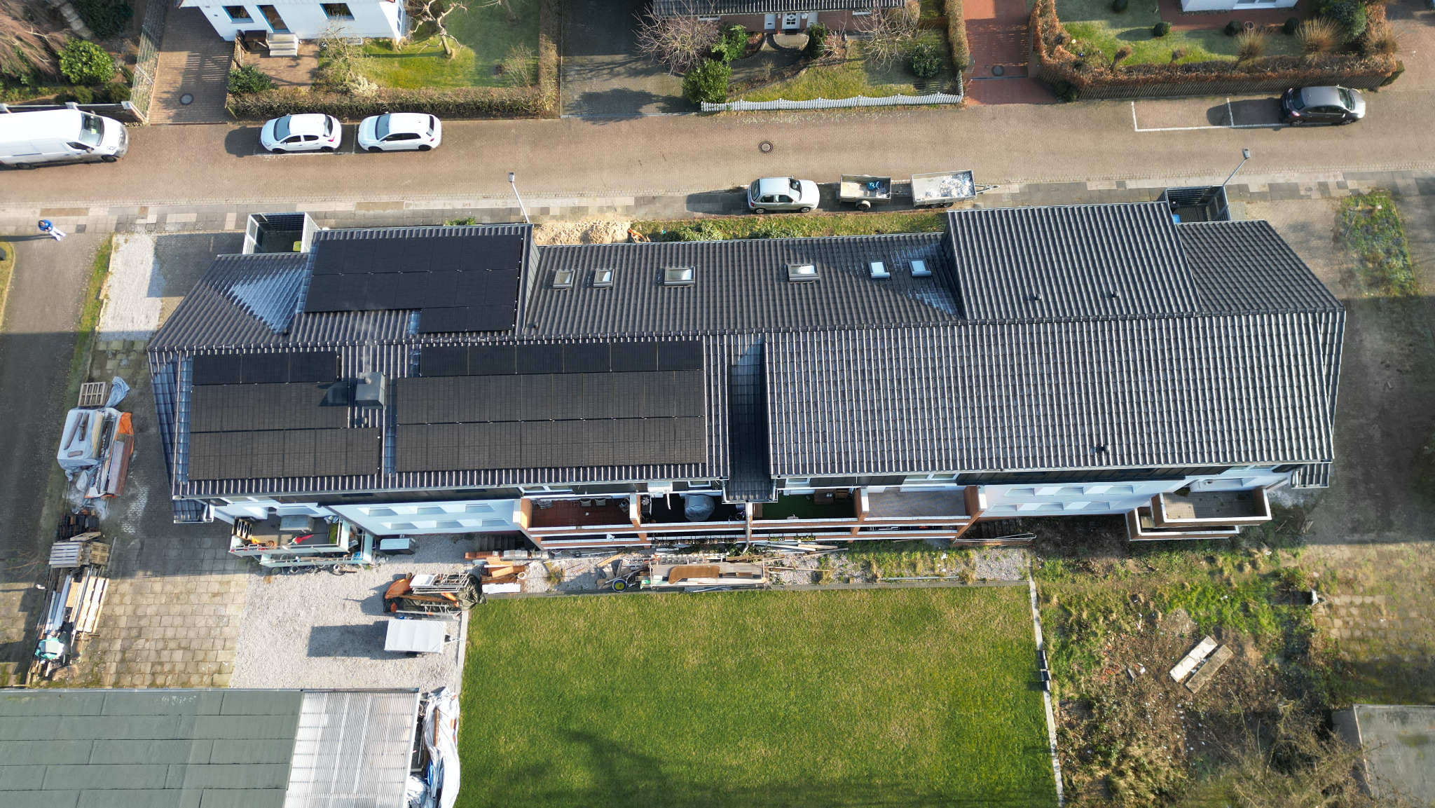 Widok z lotu ptaka na budynek z szarym dachem, na którym częściowo zamontowane są czarne panele słoneczne, widoczne okna dachowe oraz otoczenie budynku z trawnikiem i zaparkowanymi samochodami.