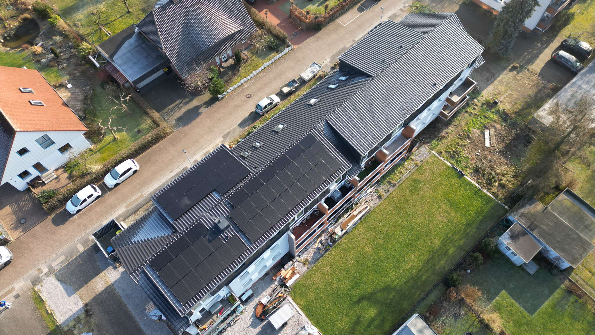 Widok z lotu ptaka na budynek z ciemną dachówką i panelami słonecznymi, otoczony zielenią i zabudową mieszkalną.