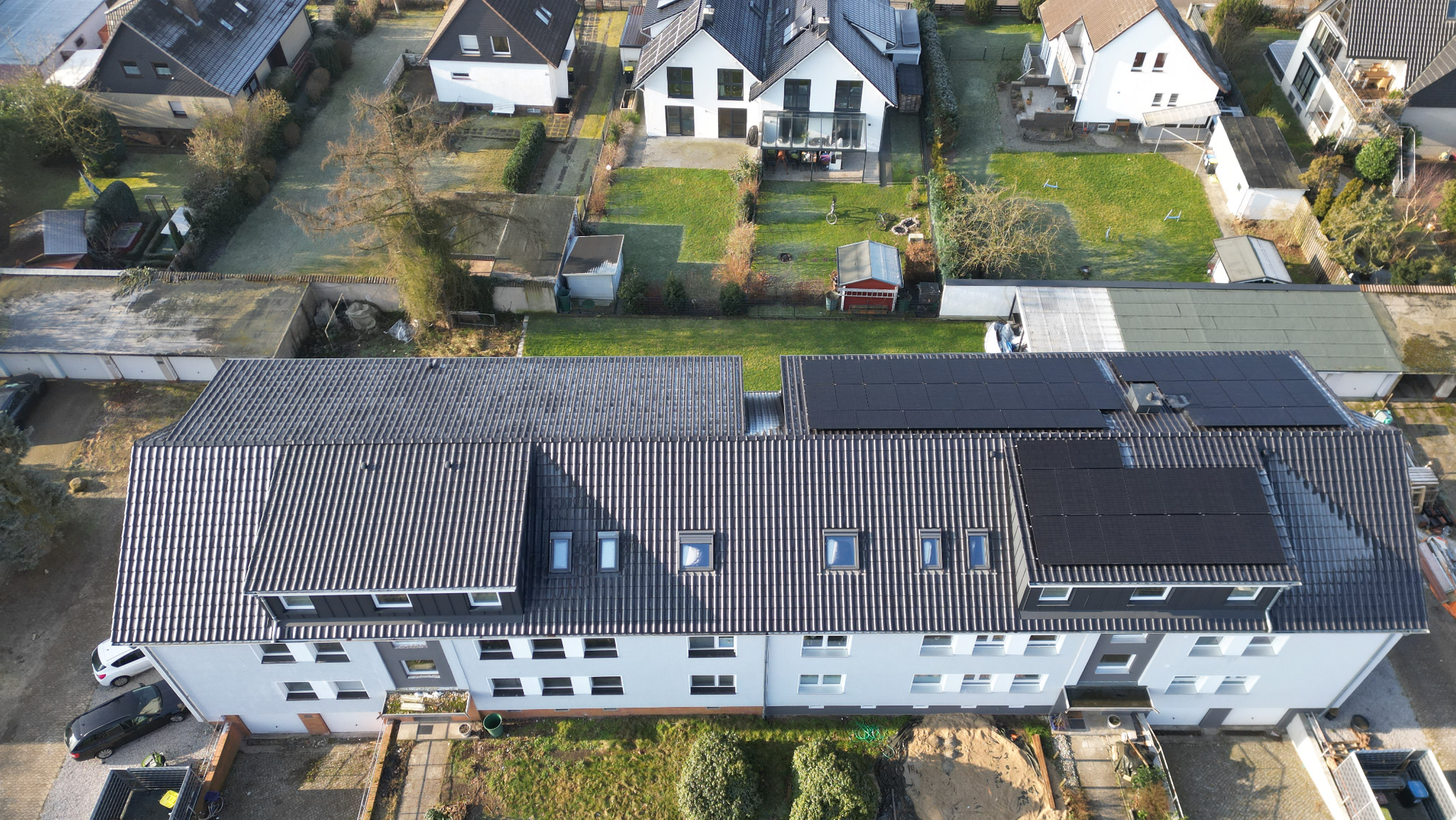 Widok z góry na budynek wielorodzinny z częściowo odnowionym dachem pokrytym ciemnoszarą dachówką karpiówką i panelami słonecznymi, otoczony zielenią i innymi budynkami.