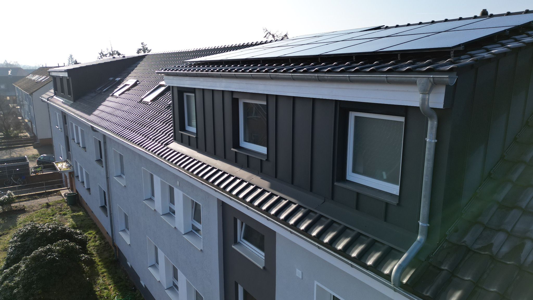 Wielorodzinny budynek z ciemnoszarą blachą na dachu i panelami słonecznymi, widok z góry na rynny i okna dachowe.