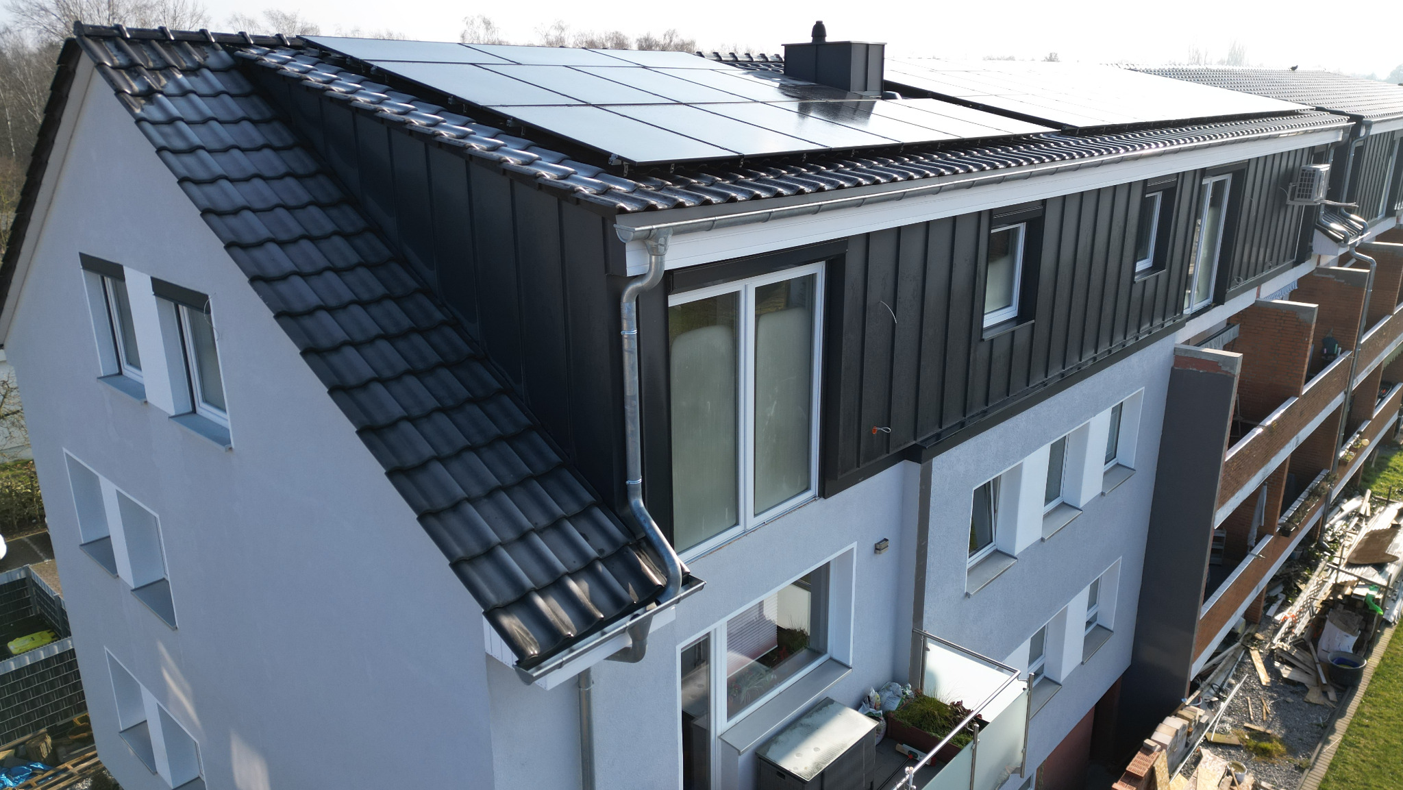 Widok z góry na budynek wielorodzinny z ciemną dachówką ceramiczną, panelami słonecznymi i obróbkami blacharskimi, z widocznymi rynnami i elementami wentylacji.