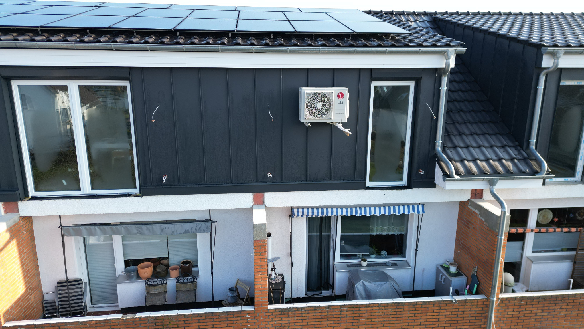 Widok z góry na dom z panelami słonecznymi na dachu, czarną blachą trapezową na ścianie, klimatyzatorem LG i rynnami.