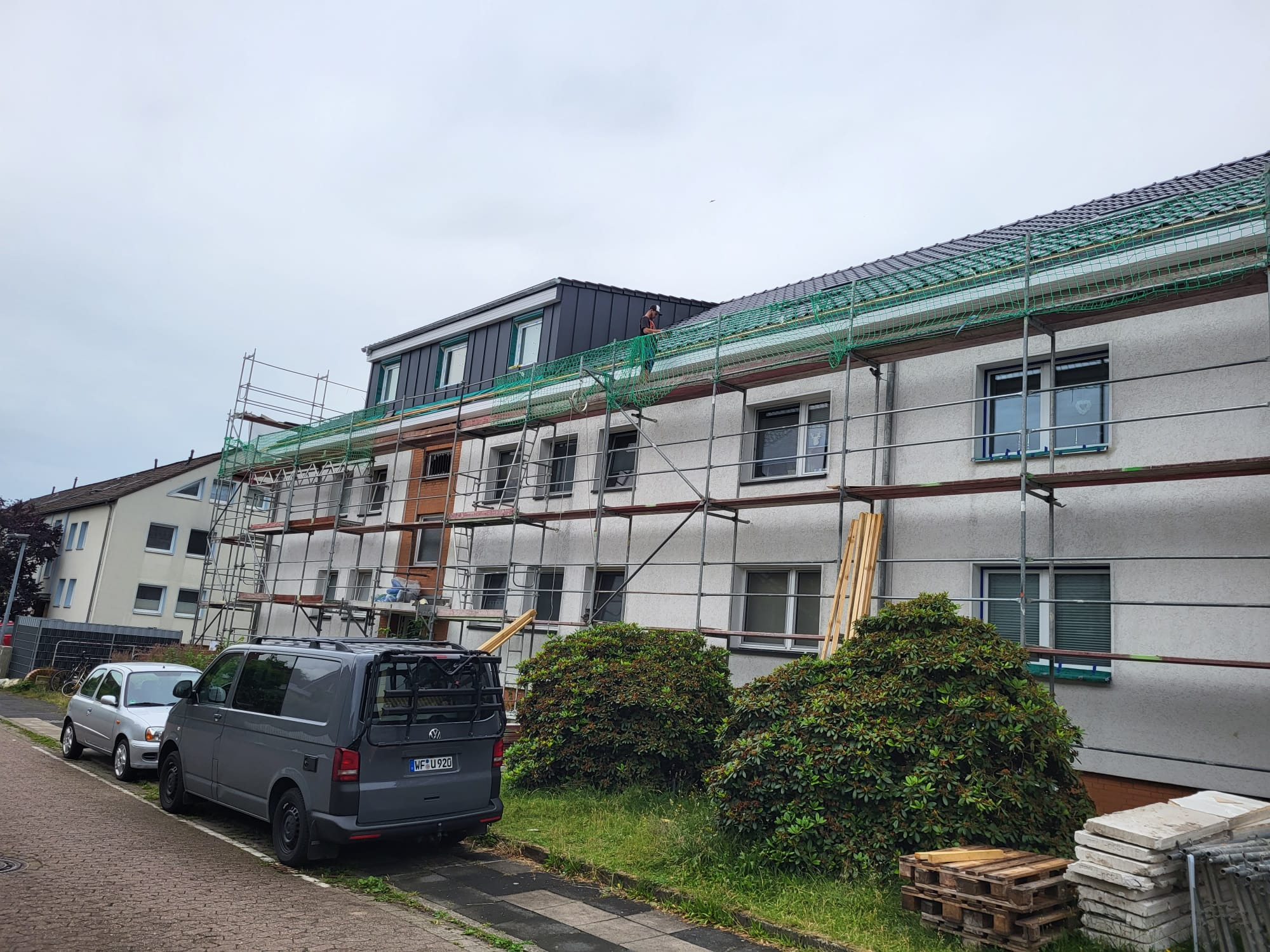 Pracownik na rusztowaniu zabezpieczonym siatką, montuje obróbki blacharskie na dachu budynku wielorodzinnego z nową, grafitową nadbudową.