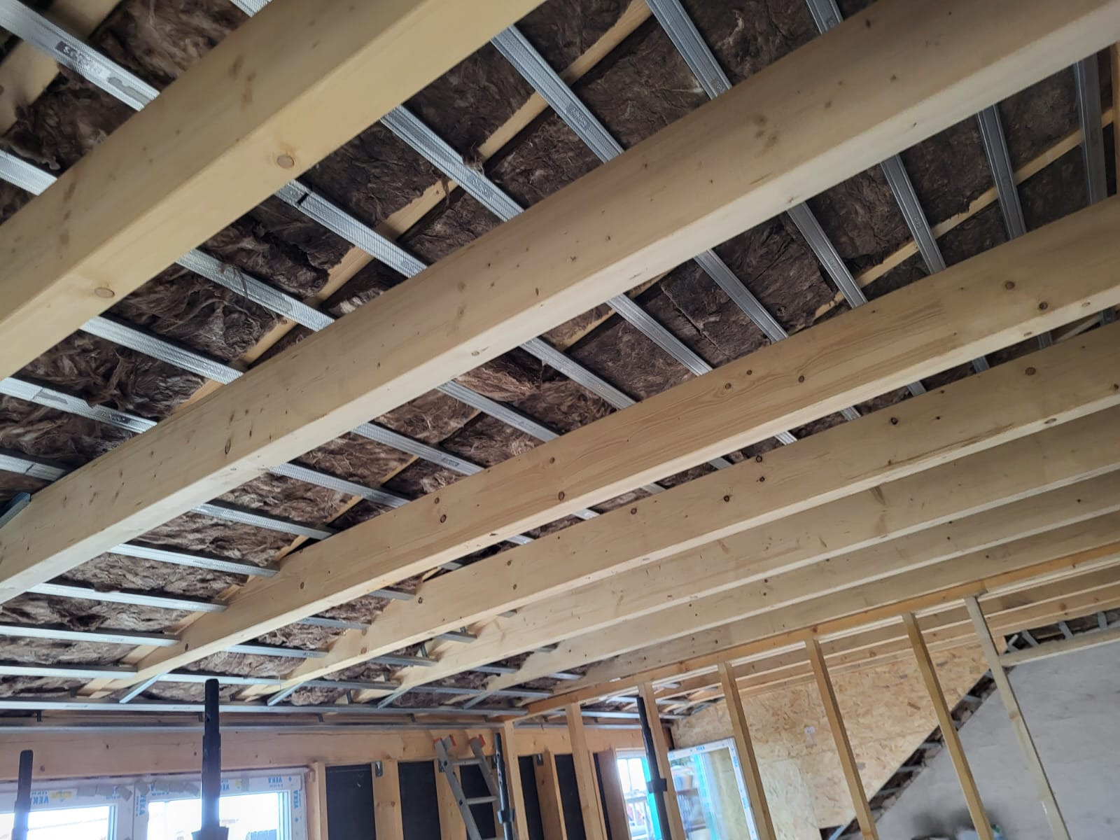 Wnętrze poddasza w trakcie ocieplania: widoczne drewniane belki stropowe, metalowe profile pod płyty gipsowe oraz brązowa wełna mineralna między nimi.