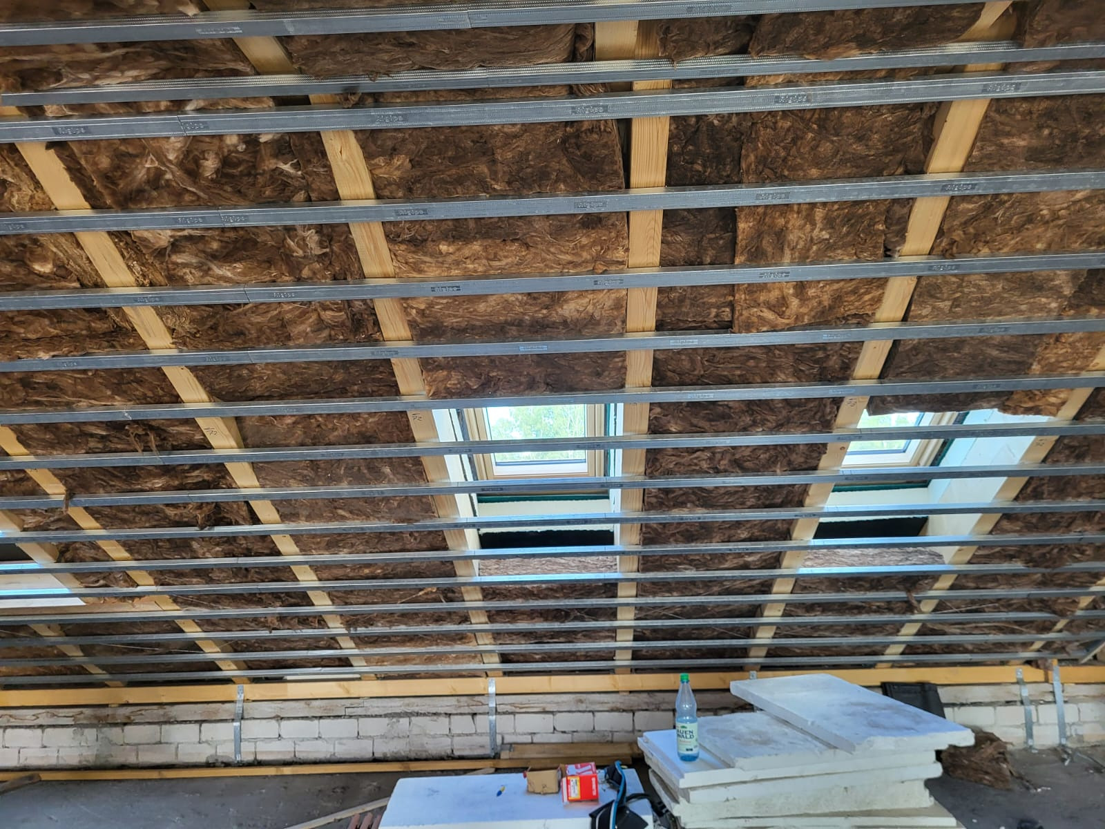 Poddasze w trakcie ocieplania wełną mineralną, widoczne metalowe profile mocujące i okna dachowe, na dole składowane płyty izolacyjne i butelka wody.