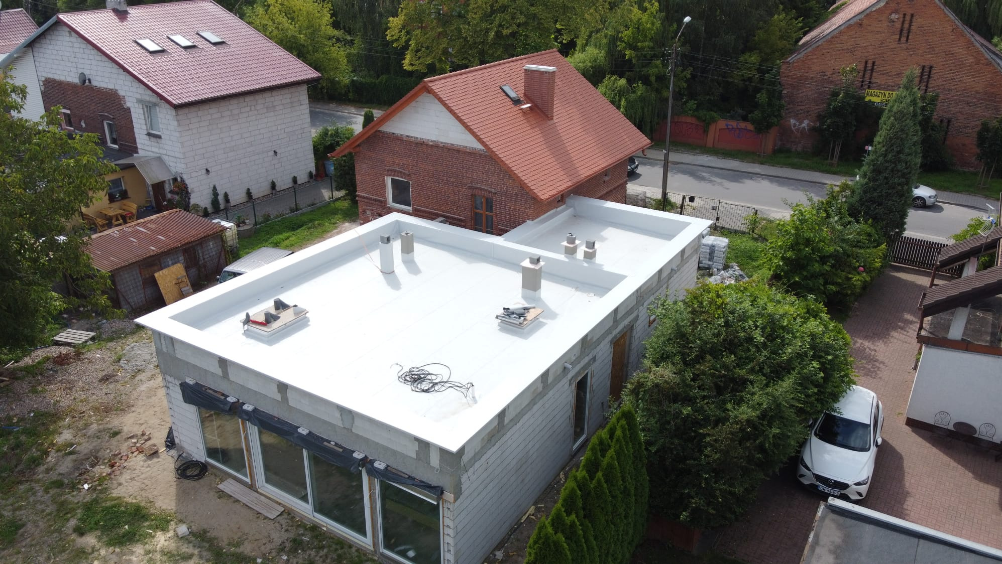 Biały, płaski dach na budynku w trakcie budowy, widok z góry, kominy wentylacyjne i narzędzia na dachu, w tle domy z dachami skośnymi.