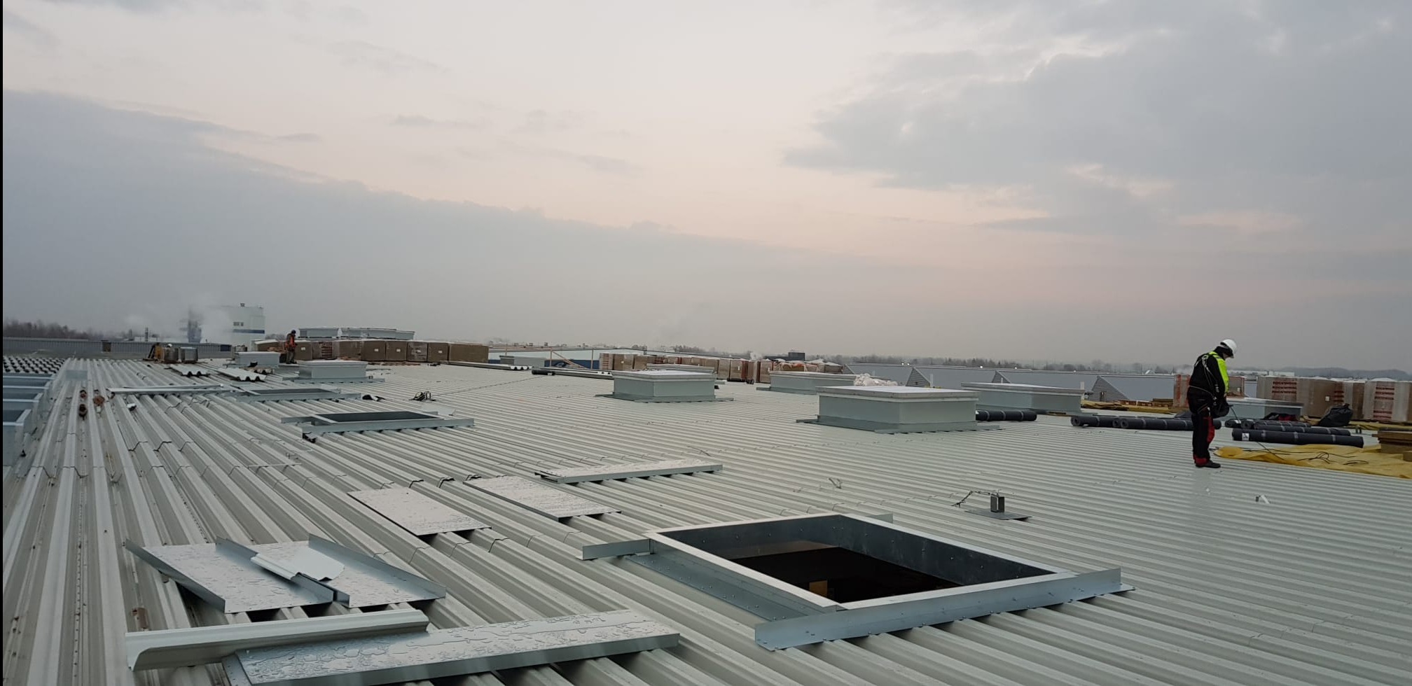 Pracownik w kasku i kamizelce odblaskowej na dachu przemysłowym z blachy trapezowej, widoczne otwory okienne, obróbki blacharskie i materiały budowlane w tle.