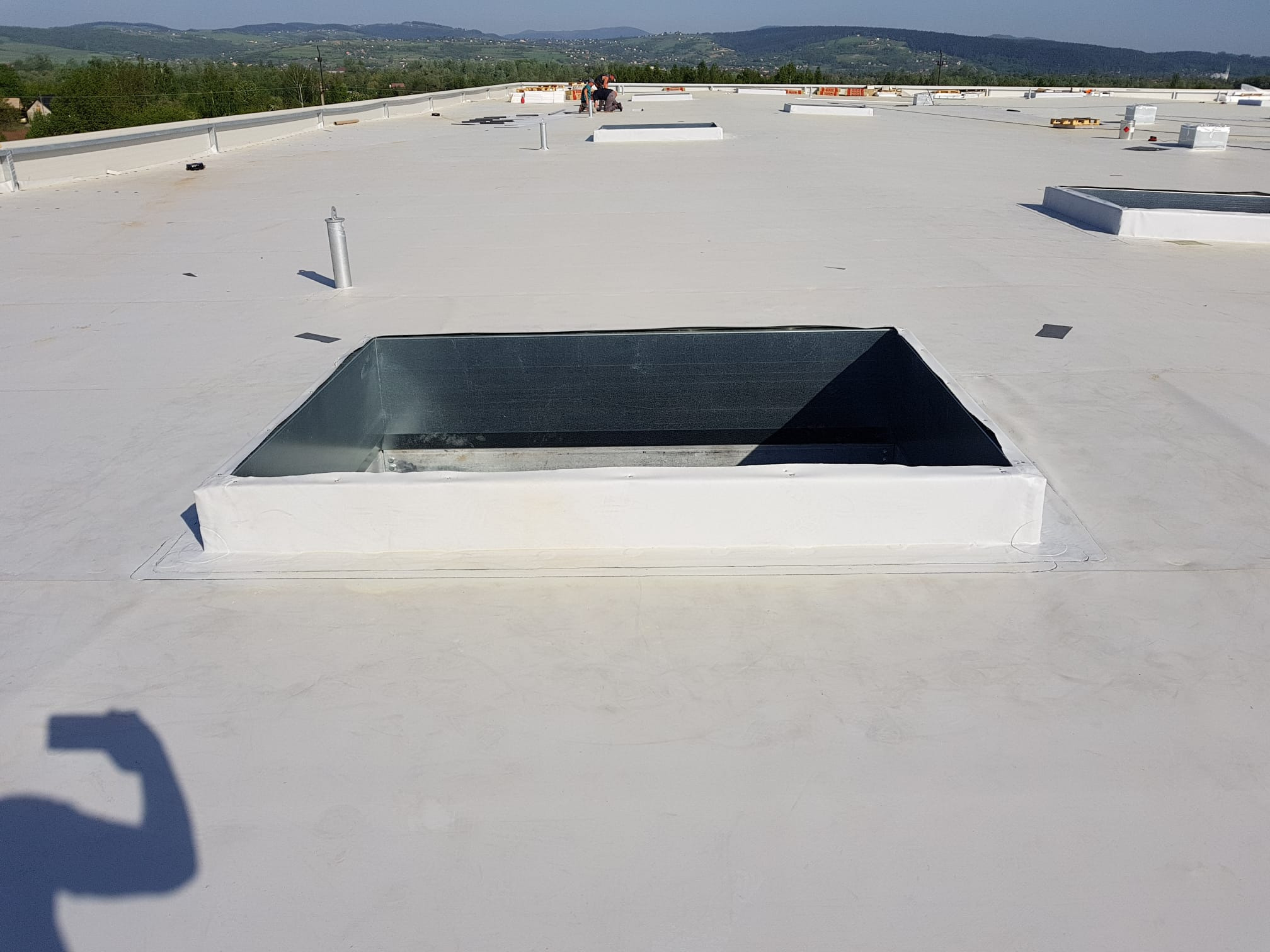 Biała, płaska powierzchnia dachu z zainstalowanymi świetlikami i kominkami wentylacyjnymi, w tle widoczni pracownicy i górzysty krajobraz.