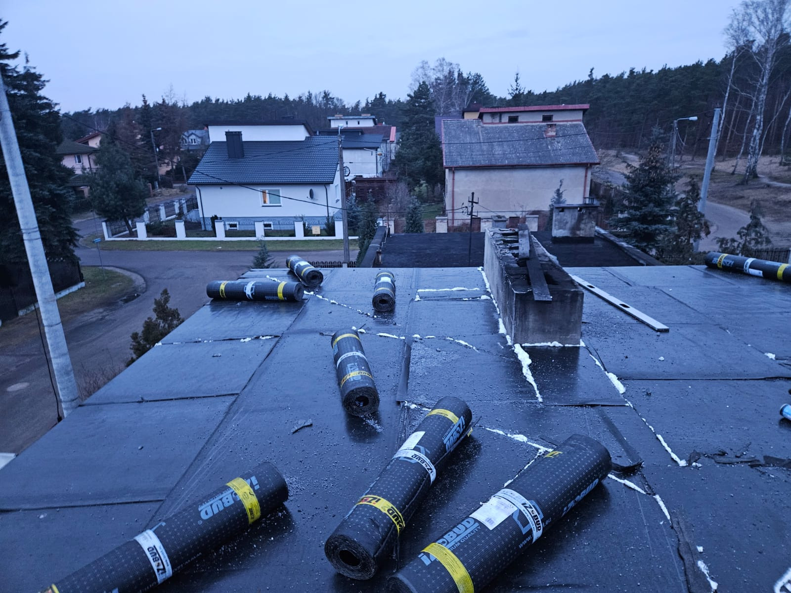 Bitumiczna papa w rolkach leży na płaskim dachu pokrytym czarnym materiałem izolacyjnym, widoczne uszczelnienia pianką montażową, w tle zabudowa jednorodzinna i las.