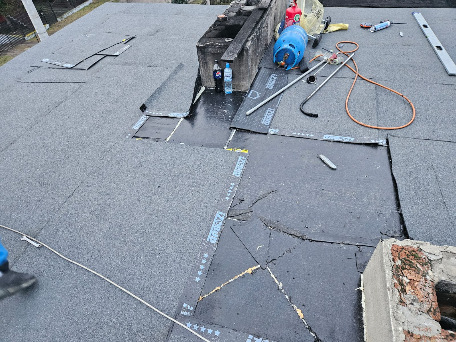 Fragment dachu płaskiego w trakcie prac, widoczne warstwy izolacji, butla gazowa, narzędzia dekarskie i komin z cegły