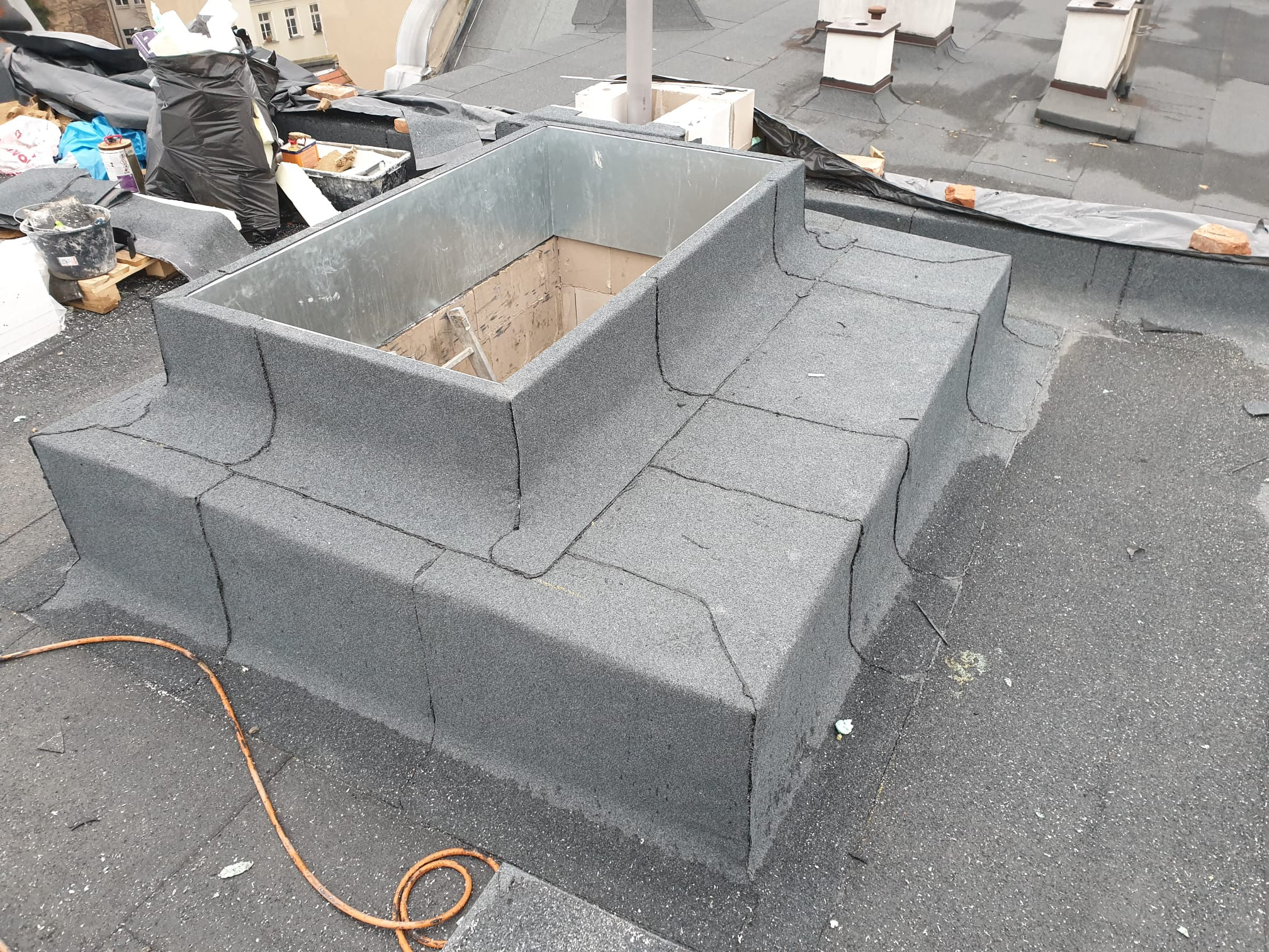 Wykończona obróbka blacharska komina z papy na dachu płaskim, widoczne narzędzia i materiały budowlane w tle.