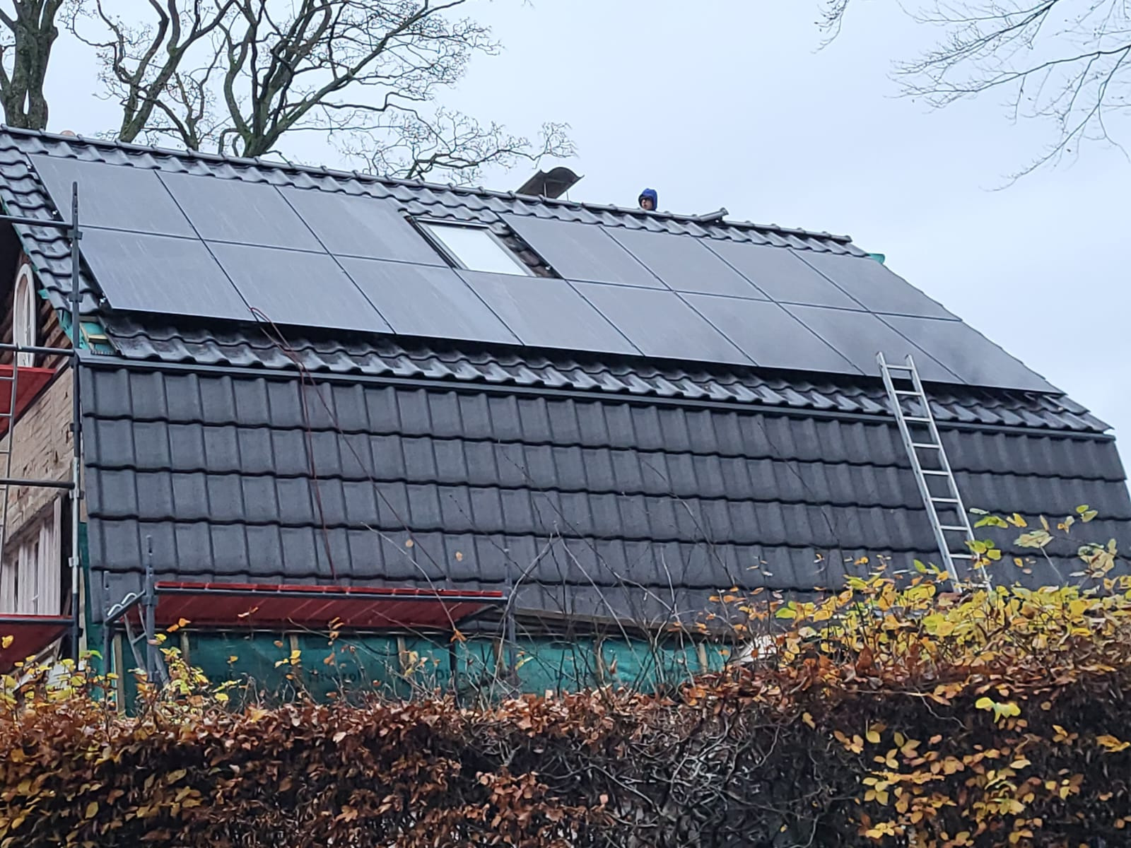 Montaż paneli fotowoltaicznych na dachu pokrytym ciemną dachówką, widoczna drabina oparta o dach i pracownik w niebieskiej czapce na szczycie.