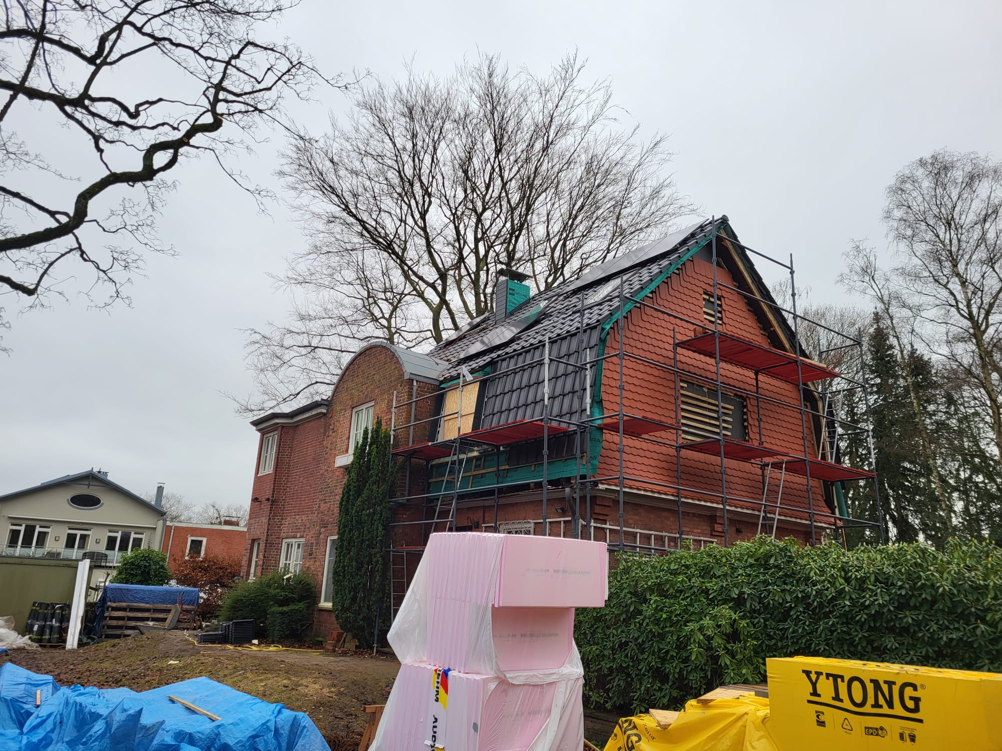 Dom z czerwonej cegły w trakcie prac dekarskich, widoczne rusztowania, częściowo pokryty dachówką, materiały izolacyjne na pierwszym planie.