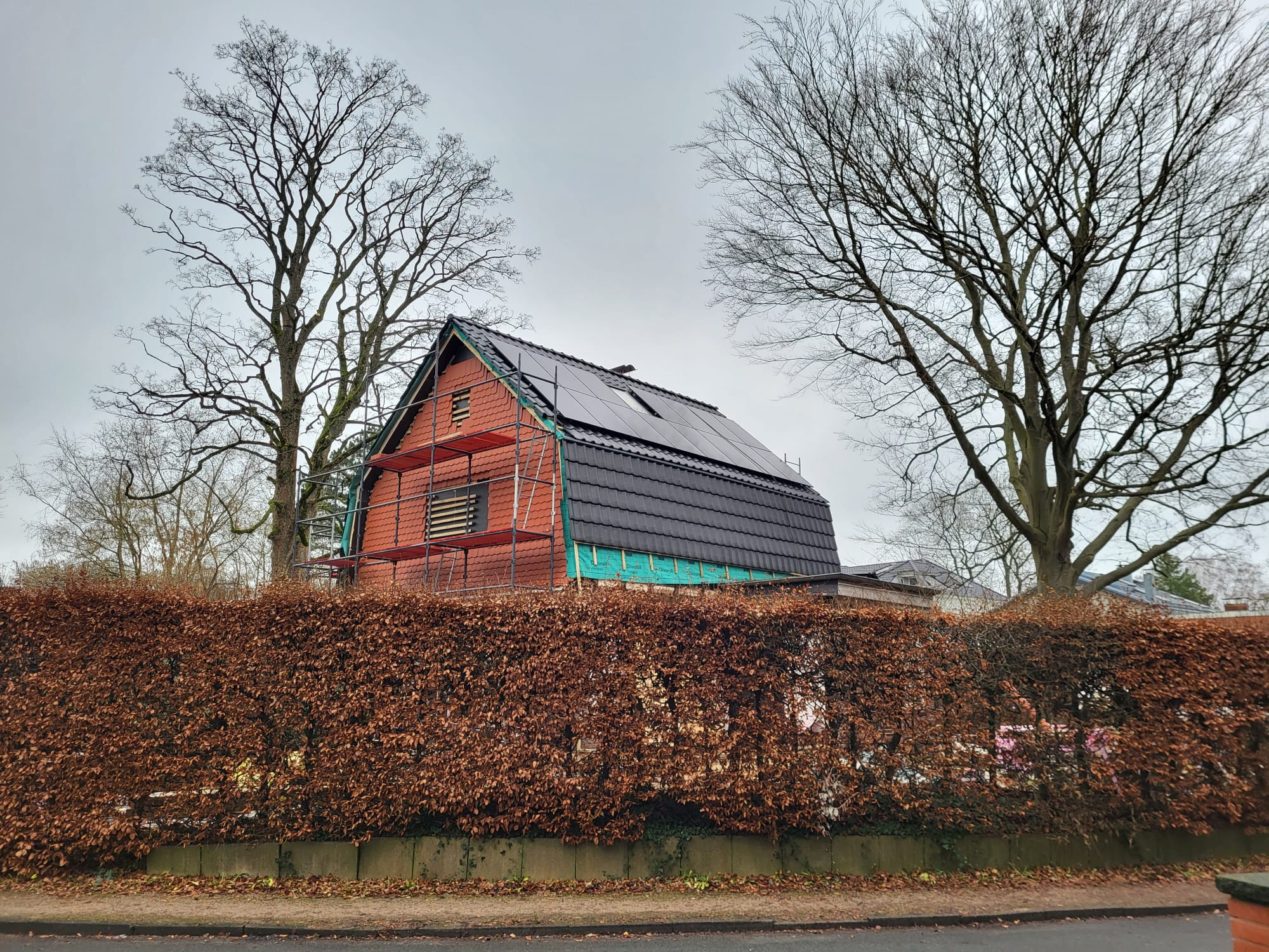 Dom w trakcie prac dekarskich, widoczne rusztowania, fragment nowej dachówki i panele słoneczne, tło stanowią drzewa bez liści i żywopłot.