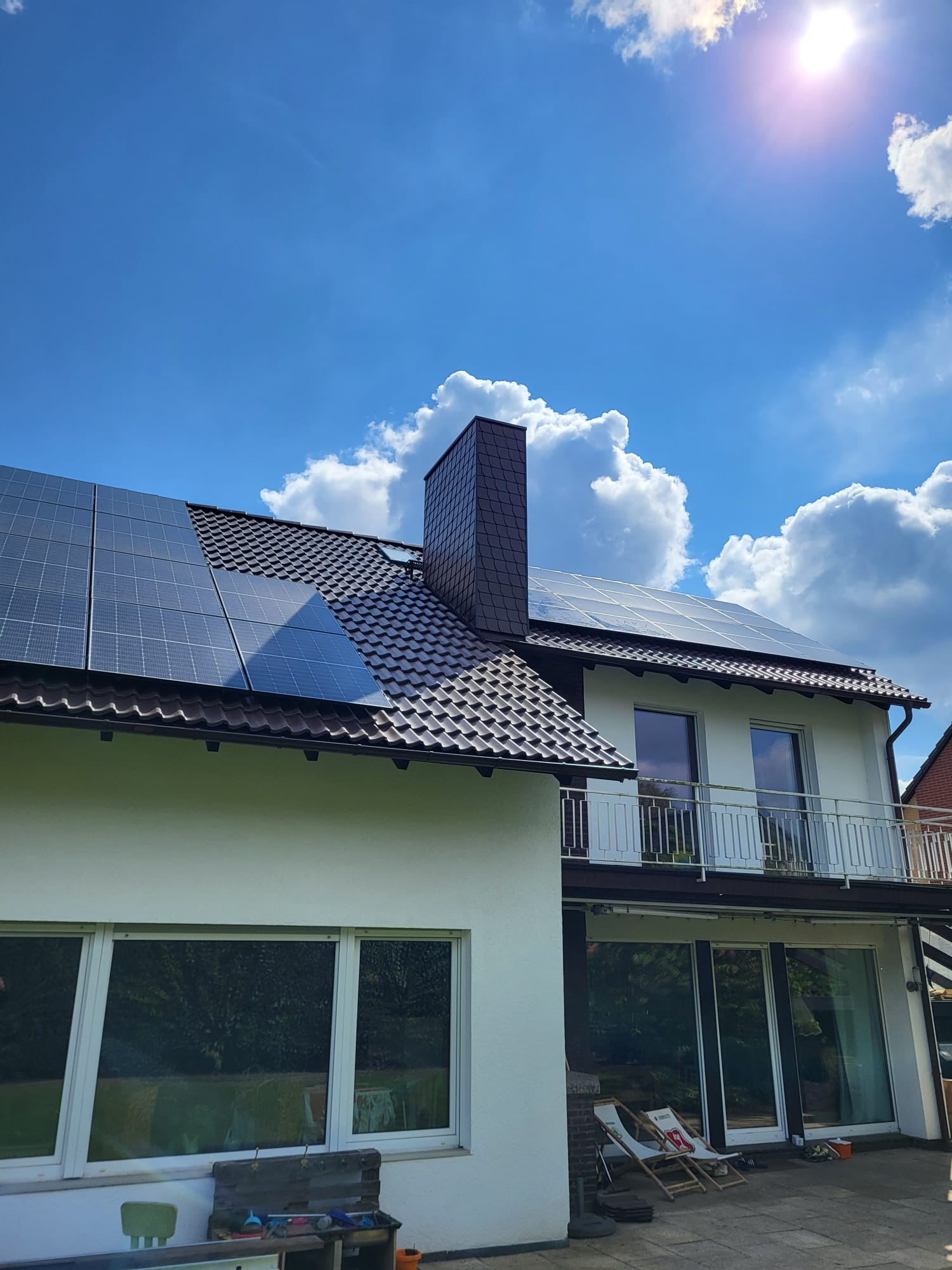 Dom z brązowym dachem i panelami słonecznymi w słoneczny dzień, widoczne białe ściany, balkon z białą balustradą i komin z ciemnej dachówki.