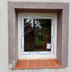 Białe okno PCV marki Kömmerling w grubym, szarym obramowaniu na elewacji. W szybie odbicie osoby robiącej zdjęcie telefonem. Parapet wyłożony czerwoną cegłą.