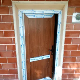 Montaż brązowych drzwi zewnętrznych z okleiną drewnopodobną, zabezpieczonych folią ochronną Kömmerling, w murze z czerwonej cegły.