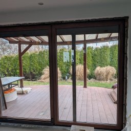 REC NOVA Wojciech Tomaszewski - Montaż brązowych drzwi przesuwnych z widokiem na taras i ogród. Widoczne surowe wykończenie ściany po montażu. Na zewnątrz pergola i meble ogrodowe.