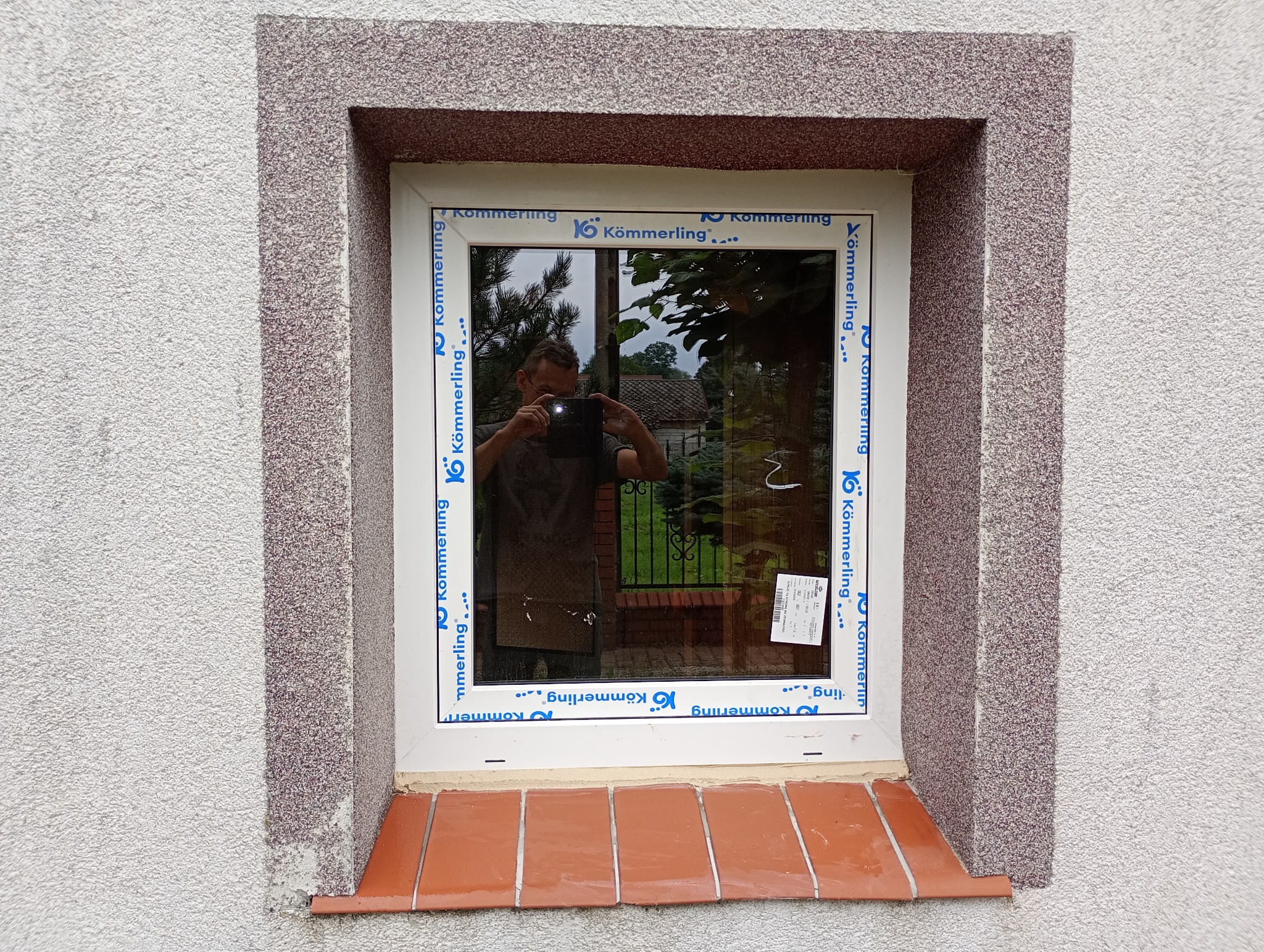 Białe okno PCV marki Kömmerling w grubym, szarym obramowaniu na elewacji. W szybie odbicie osoby robiącej zdjęcie telefonem. Parapet wyłożony czerwoną cegłą.