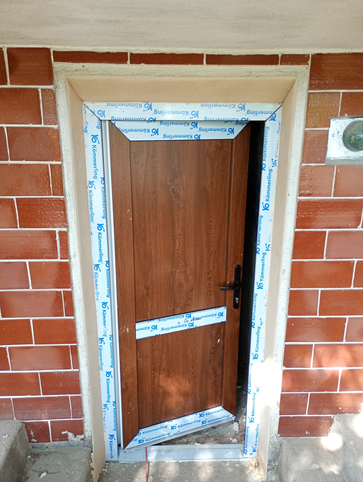 Montaż brązowych drzwi zewnętrznych z okleiną drewnopodobną, zabezpieczonych folią ochronną Kömmerling, w murze z czerwonej cegły.