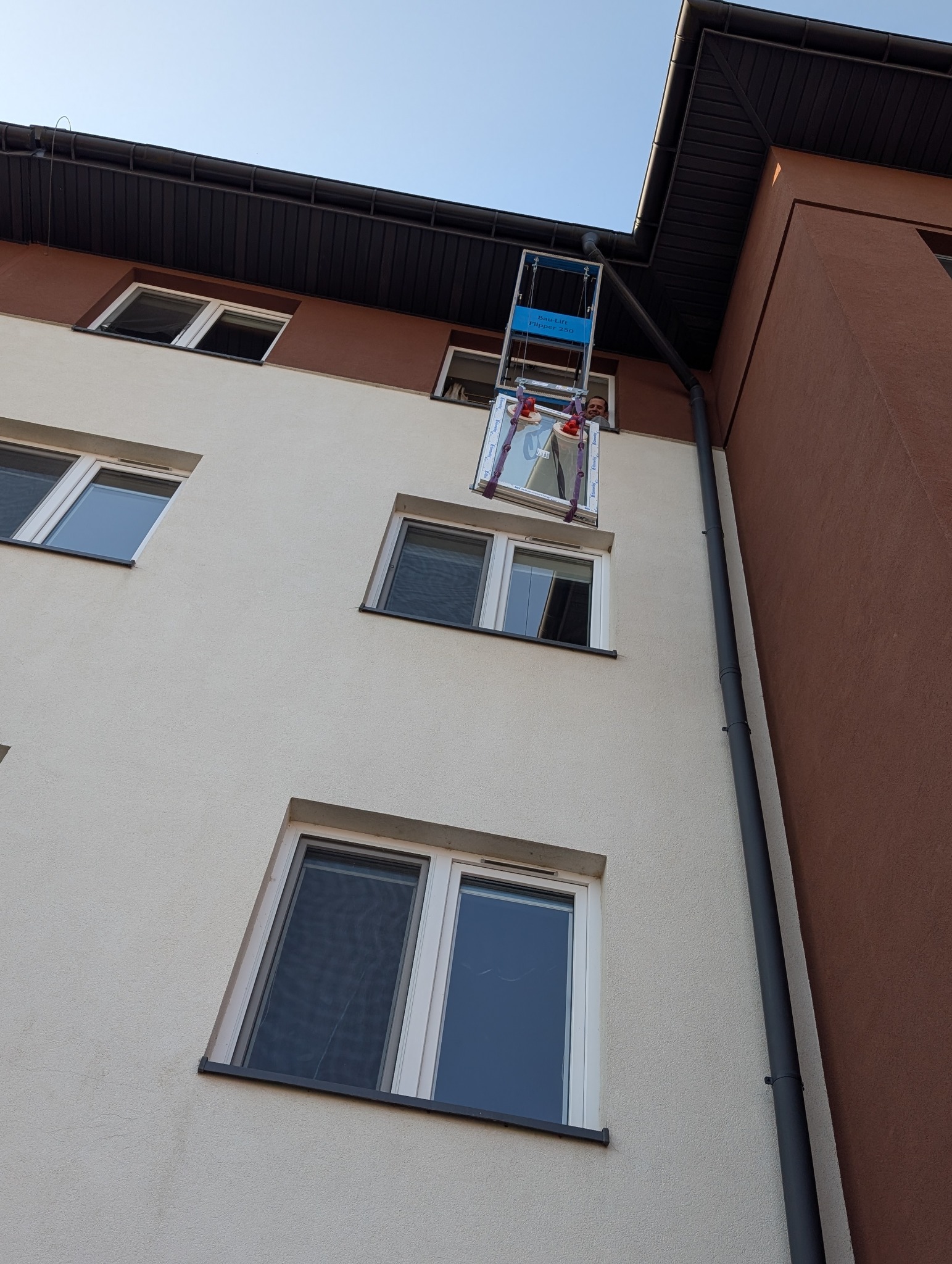 Montaż okna PCV na wysokości za pomocą podnośnika. Widoczny pracownik i okno w trakcie instalacji na tle elewacji budynku.