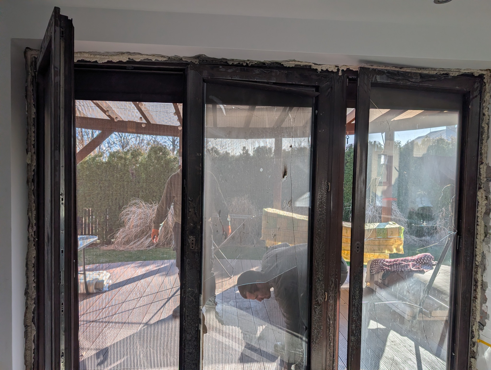 Demontaż okna balkonowego z uszkodzoną szybą. Widoczne ślady pianki montażowej i pracownik w trakcie demontażu. Na zewnątrz taras i zieleń.