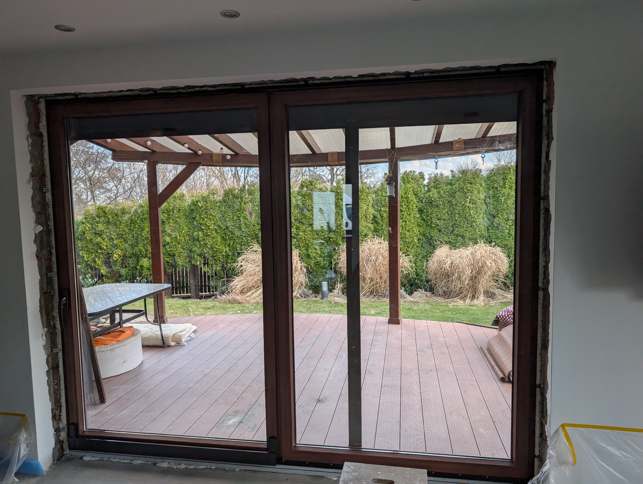 Montaż brązowych drzwi przesuwnych z widokiem na taras i ogród. Widoczne surowe wykończenie ściany po montażu. Na zewnątrz pergola i meble ogrodowe.