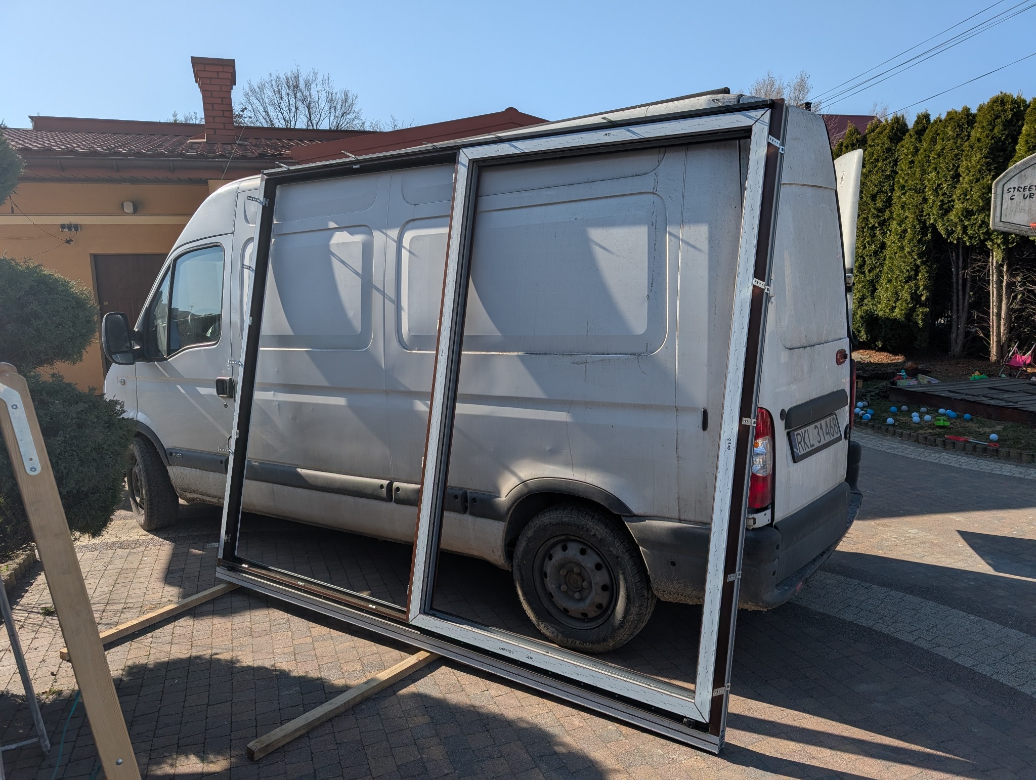 Biała rama okienna oparta o biały samochód dostawczy na zewnątrz. Widoczne elementy montażowe i drewniane podpory. W tle dom i drzewa.