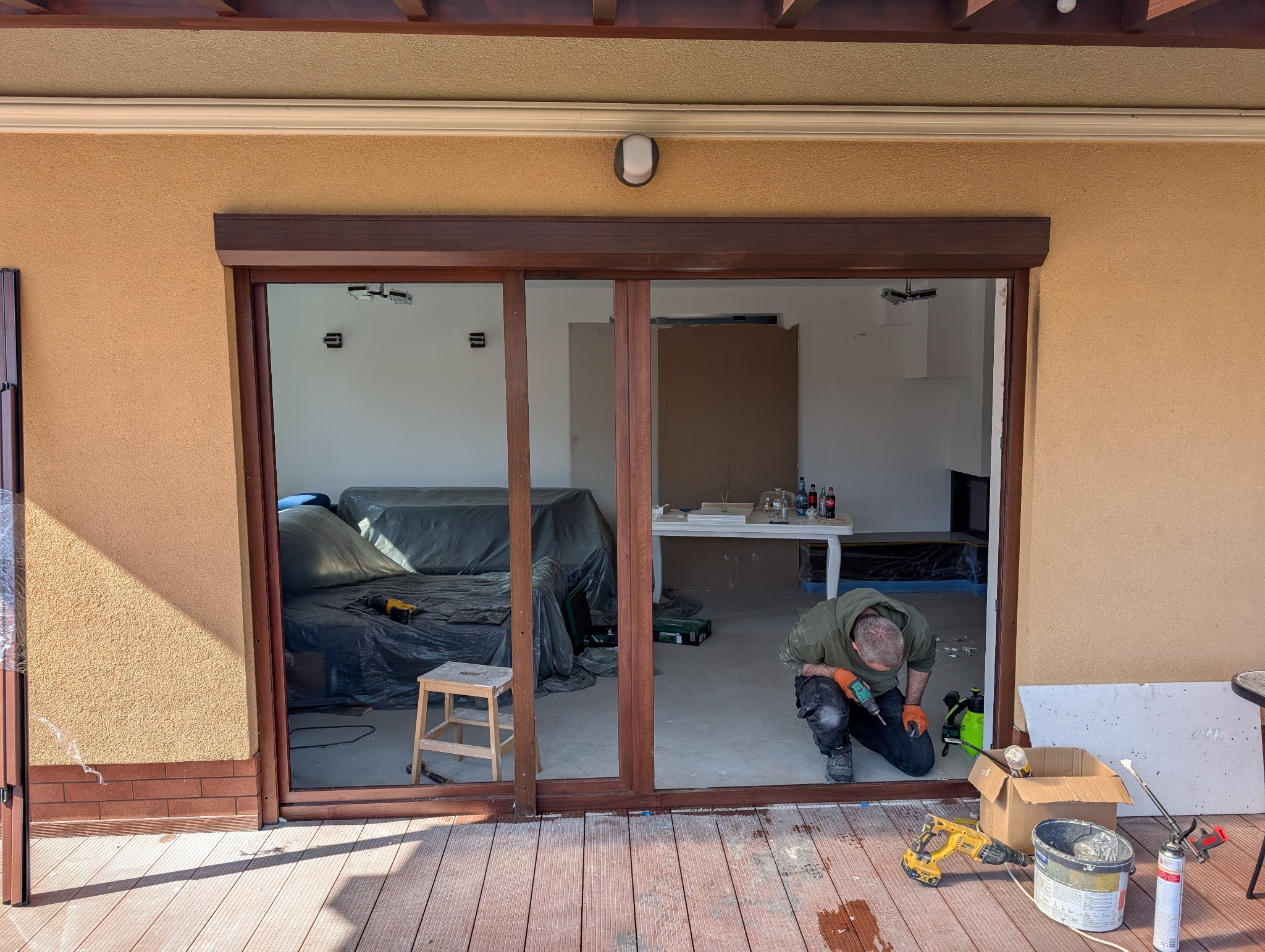 Montaż przesuwnych drzwi balkonowych z brązowymi ramami. Widoczny monter przy pracy, narzędzia i częściowo zakryte meble wewnątrz pomieszczenia.