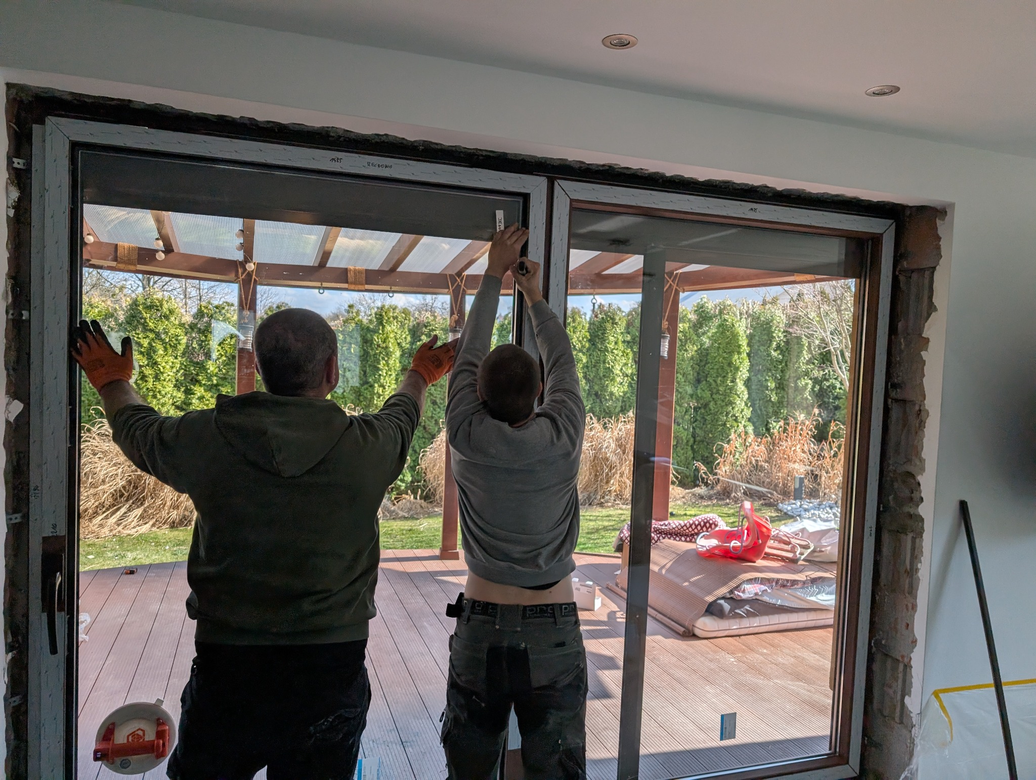 Montaż dwuskrzydłowych drzwi balkonowych z widokiem na taras i ogród. Dwóch monterów w trakcie pracy, widoczne narzędzia i elementy montażowe.