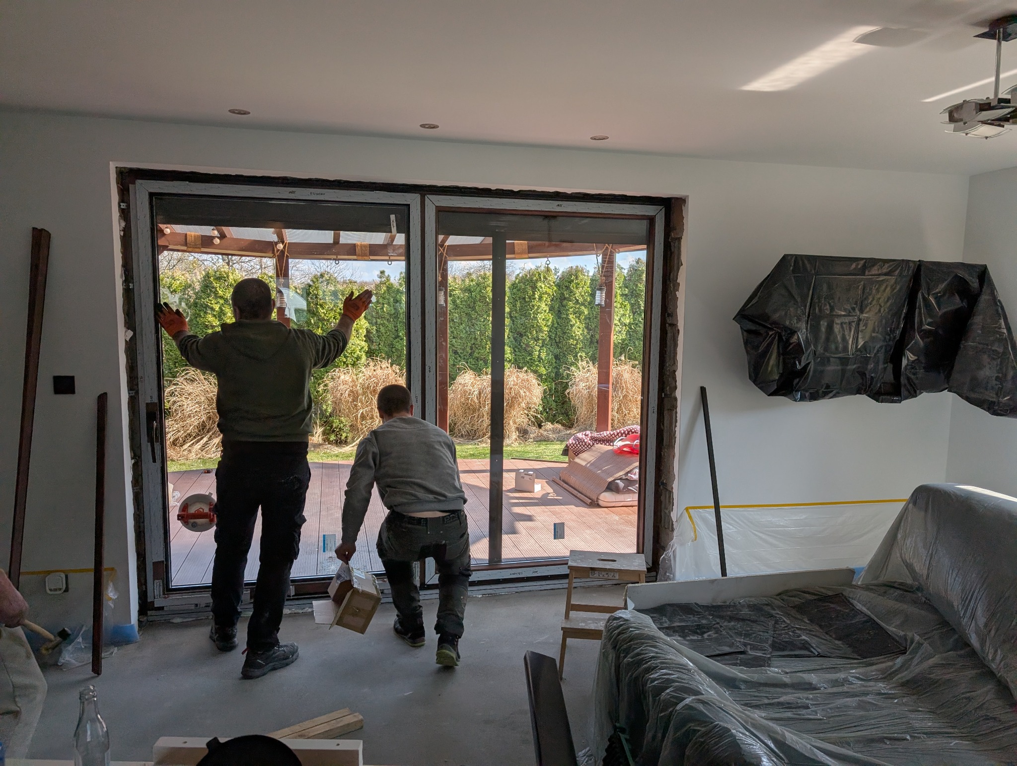 Montaż dwuskrzydłowych okien tarasowych z widokiem na ogród. Dwóch monterów podczas pracy, zabezpieczone meble folią malarską. Widoczne narzędzia i akcesoria montażowe.