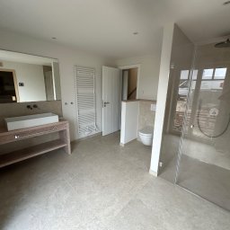 Hydro-Fliz - Jasna łazienka z beżowymi płytkami, drewnianą szafką pod umywalką i kabiną prysznicową ze szklanymi drzwiami. W lustrze odbija się osoba robiąca zdjęcie.