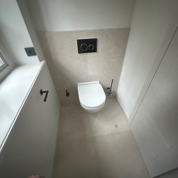 Hydro-Fliz - Nowoczesna łazienka z podwieszaną toaletą, beżowymi płytkami i czarnymi detalami: przycisk spłuczki, uchwyt na papier i szczotka toaletowa. Minimalistyczny design.