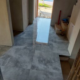 Hydro-Fliz - Ułożone, szare, matowe płytki podłogowe w korytarzu domu jednorodzinnego. Widoczny poziomica, wiertarka i częściowo ułożone schody.