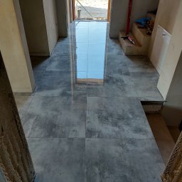 Hydro-Fliz - Ułożona podłoga z szarych, matowych płytek w korytarzu, z odbiciem nieba w połysku. Widoczne narzędzia pracy i poziomica. W tle otwarte drzwi wejściowe.