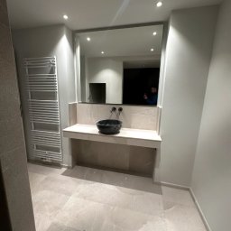 Hydro-Fliz - Wykończona łazienka w jasnych odcieniach beżu z czarną umywalką na blacie. Na ścianie grzejnik drabinkowy i duże lustro. Widoczne płytki podłogowe i oświetlenie punktowe.