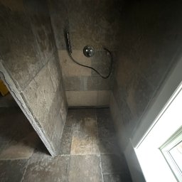 Hydro-Fliz - Wykończona kabina prysznicowa z szarymi płytkami imitującymi kamień, widoczny odpływ liniowy i bateria prysznicowa z wężem. U dołu kadru stopy osoby fotografującej.